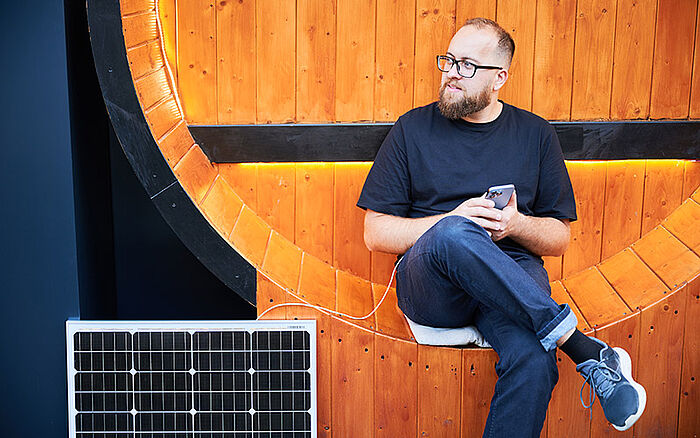 Bearded man using a smartphone connected to a photovoltaic solar panel outdoors, demonstrating practical use of solar energy for charging devices.