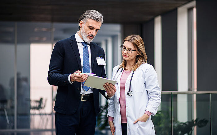 Pharmaceutical sales representative discussing medication options with a doctor while holding a tablet in a medical office.