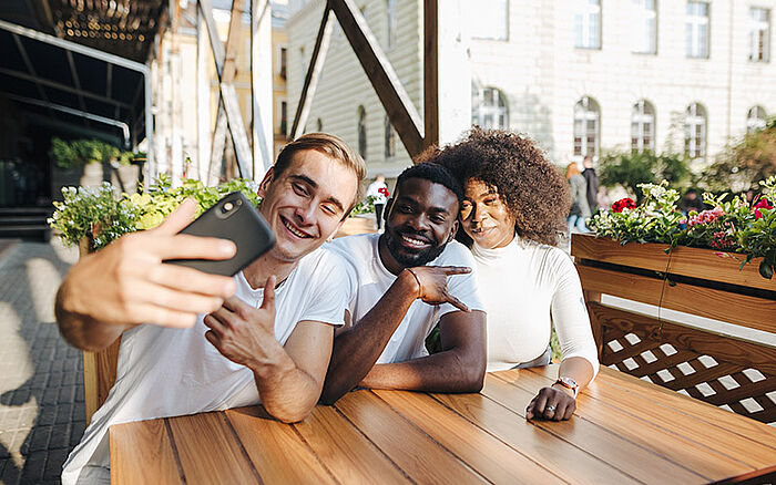  Group of intercultural friends sitting at a restaurant table, smiling and taking a selfie together.