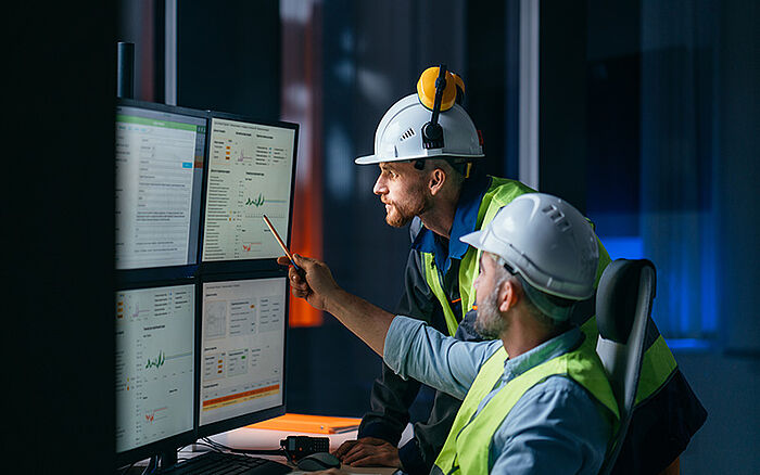 Two engineers monitor a factory assembly line using a digital SCADA-style interface, with industrial machines and data overlays in a modern manufacturing setting.