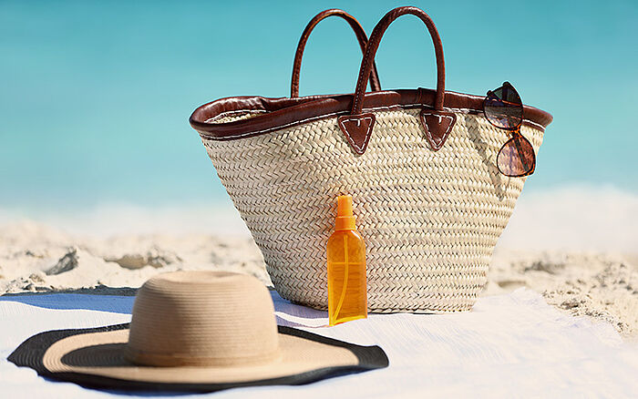 Women&rsquo;s beach accessories including a straw tote bag, sun hat, and sunscreen bottle on sand with a blue ocean in the background.