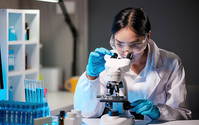 Asian female scientist examining a sample under a microscope in a high-tech pharmaceutical lab, with petri dishes and research equipment in the background.