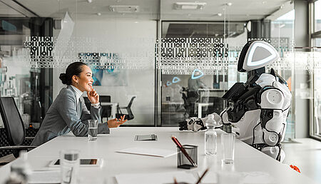 Side view of a smiling Asian businesswoman engaging in a conversation with a robot in a modern conference hall, emphasizing a professional and futuristic atmosphere.