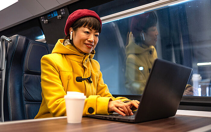 Woman working on a laptop while traveling by train, seated by the window with natural light illuminating her workspace.