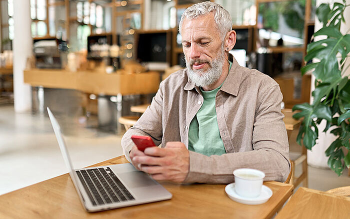 Middle-aged gray-haired businessman sitting at a caf&eacute; table, holding a smartphone and looking at the screen while working or texting.